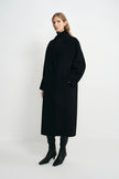 A woman modeling a long black double-breasted coat with a high black turtleneck, paired with black pointed-toe ankle boots, against a plain light gray studio background.