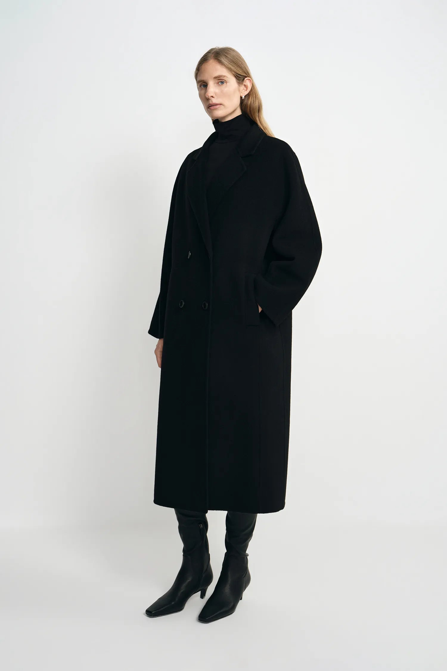 A woman modeling a long black double-breasted coat with a high black turtleneck, paired with black pointed-toe ankle boots, against a plain light gray studio background.