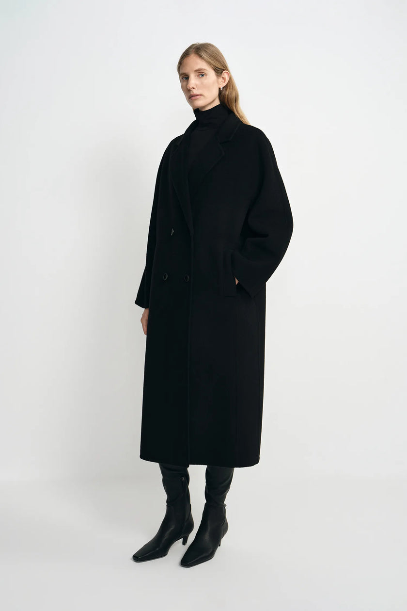 A woman modeling a long black double-breasted coat with a high black turtleneck, paired with black pointed-toe ankle boots, against a plain light gray studio background.