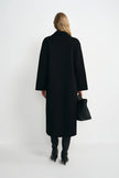 A woman shown from the back wearing a long black coat, black boots, and holding a black handbag against a plain light gray studio background.