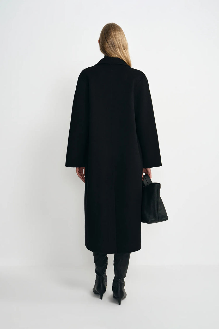 A woman shown from the back wearing a long black coat, black boots, and holding a black handbag against a plain light gray studio background.