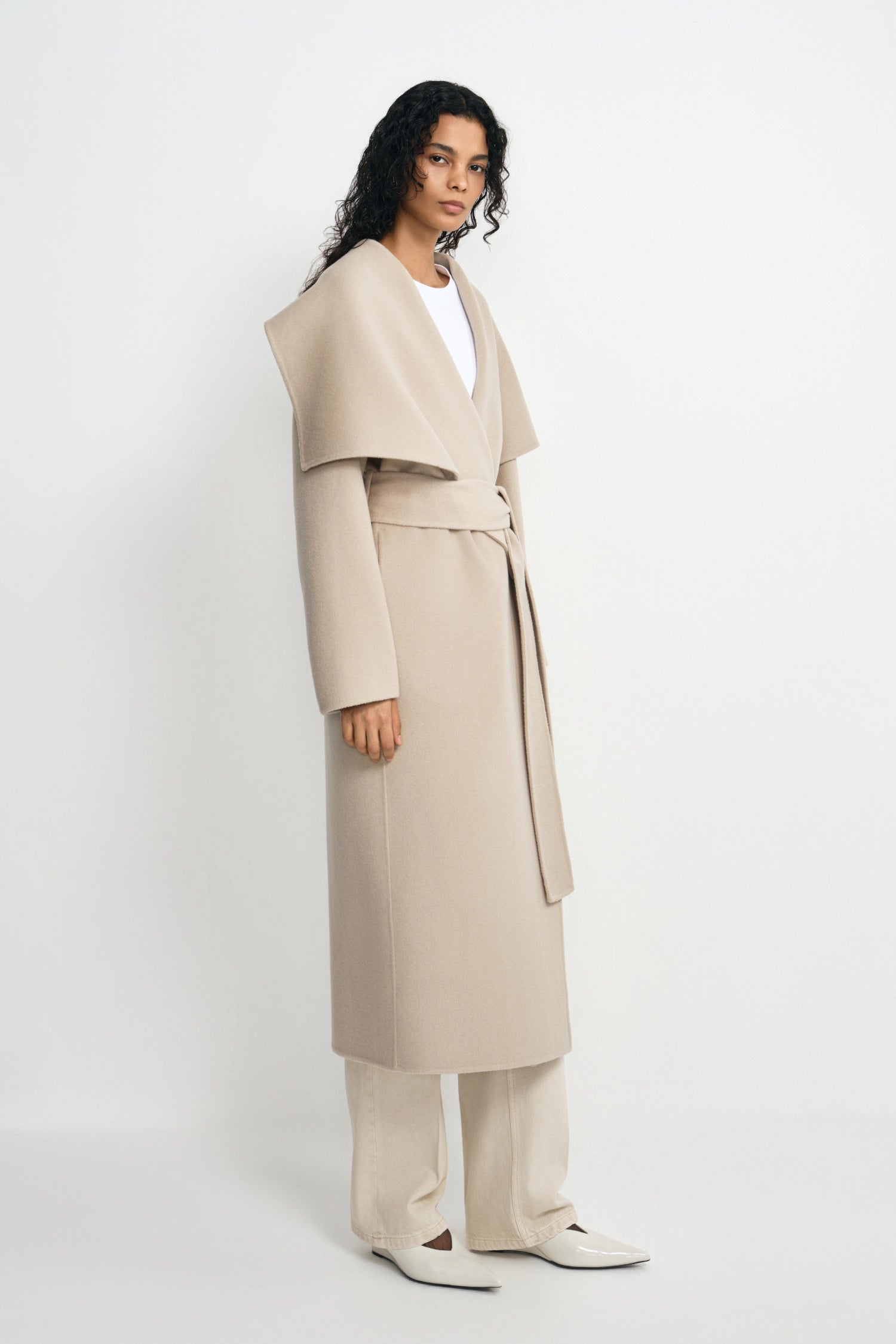 A model stands in a studio wearing a long, draped beige wrap coat with a wide collar and waist tie over a white top and matching beige trousers.