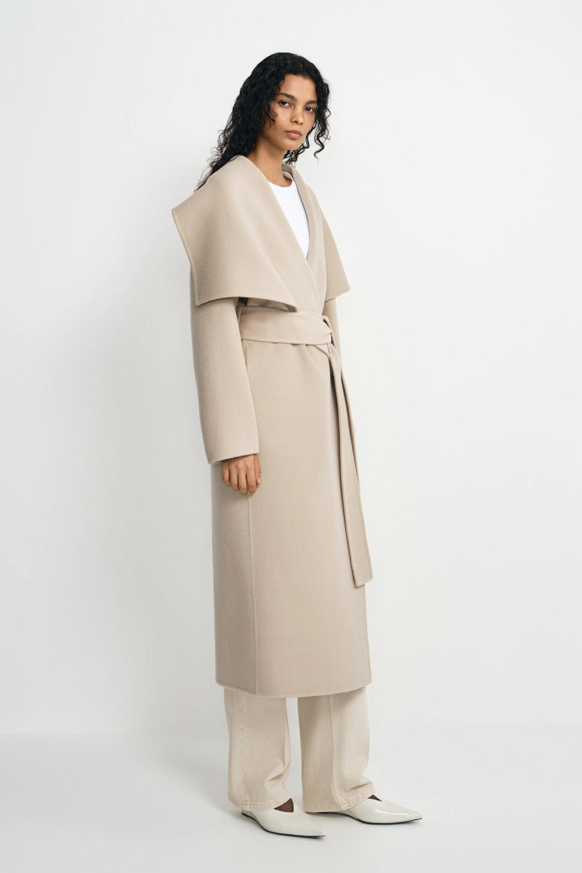 A model stands in a studio wearing a long, draped beige wrap coat with a wide collar and waist tie over a white top and matching beige trousers.