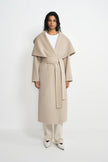A model stands against a plain white studio background wearing a long, light beige wrap coat with a wide shawl collar and self-tie belt, styled over a white top, cream trousers, and white pointed shoes.
