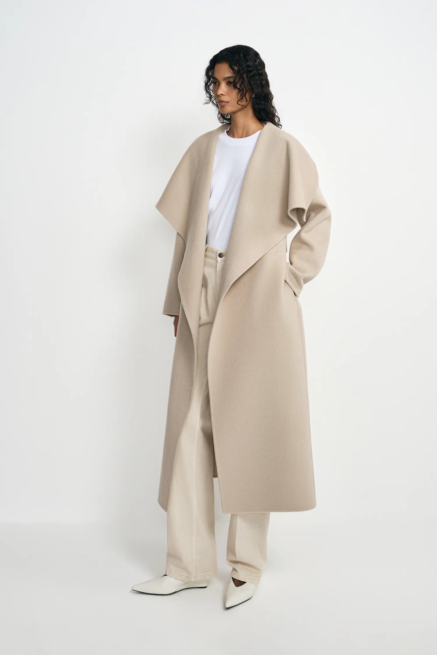 A model stands in a studio wearing a long, oversized beige wrap coat over a white top and matching beige wide-leg trousers, paired with white pointed shoes.