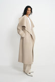 A model wearing a long, wide-collar beige coat over a white top and matching beige trousers, standing against a clean white studio background.