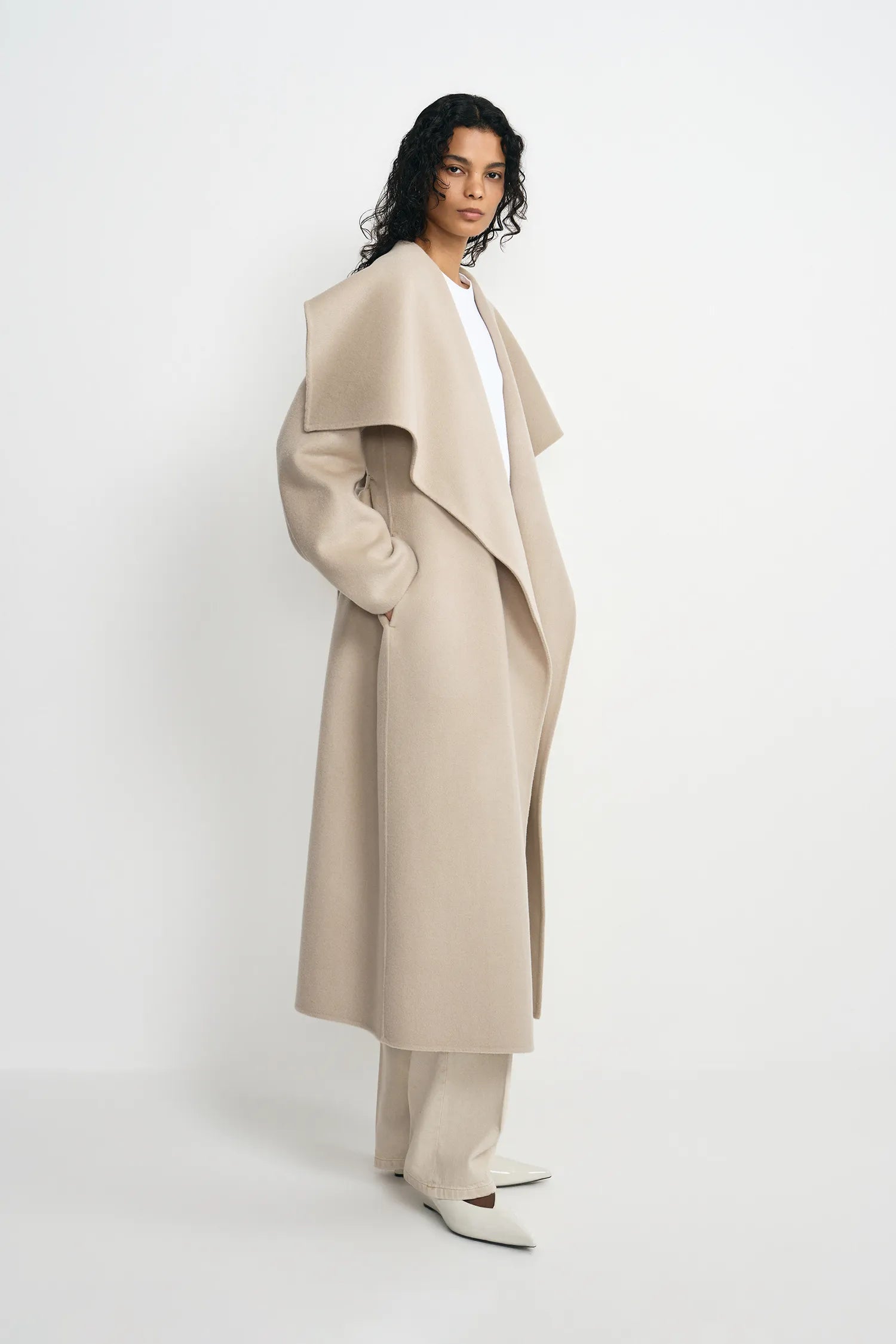 A model wearing a long, wide-collar beige coat over a white top and matching beige trousers, standing against a clean white studio background.