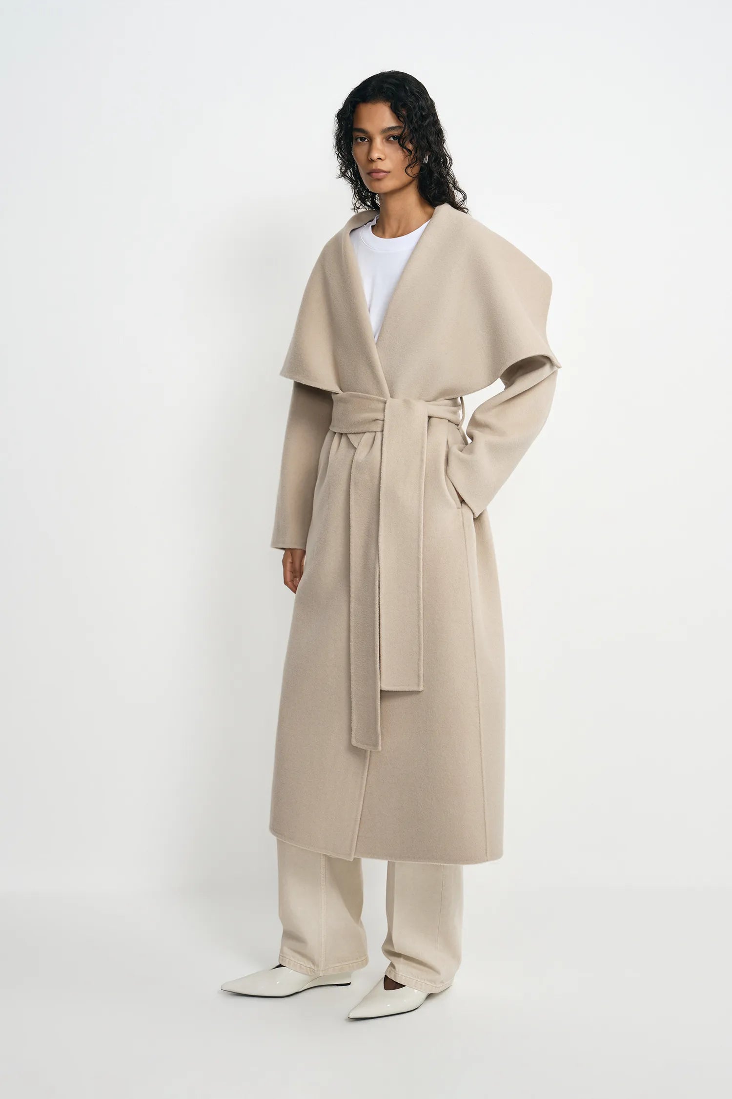 A model stands against a plain white studio background wearing a long, oversized beige wrap coat with a self-tie belt, matching beige trousers, and white pointed flats.