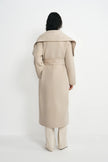 A model shown from the back wearing a long beige belted coat with dramatic shoulder folds, paired with matching beige trousers, against a plain white studio background.