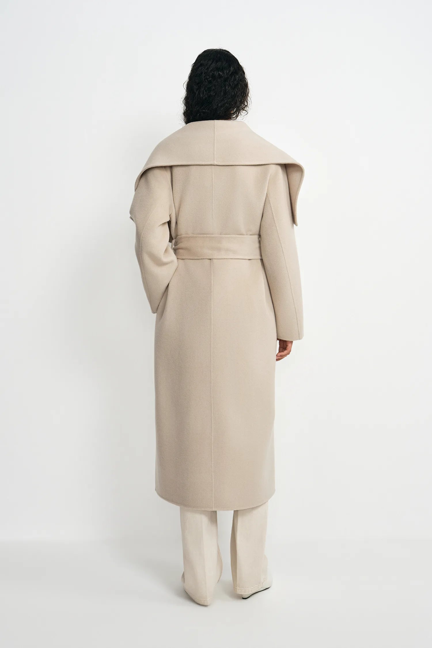 A model shown from the back wearing a long beige belted coat with dramatic shoulder folds, paired with matching beige trousers, against a plain white studio background.
