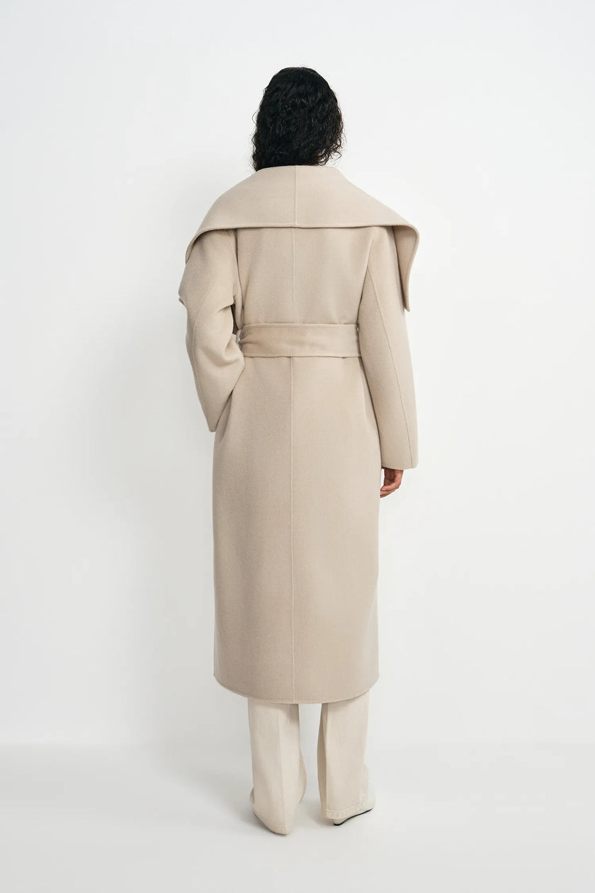 A model shown from the back wearing a long beige belted coat with dramatic shoulder folds, paired with matching beige trousers, against a plain white studio background.
