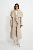 A model stands against a plain white studio background wearing a long, light beige wrap coat with a wide shawl collar and self-tie belt, styled over a white top, cream trousers, and white pointed shoes.