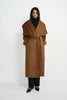 A model stands against a plain light gray studio background wearing a long camel-brown wrap coat with a wide shawl collar, a matching waist tie, black pants, and black pointed flats.