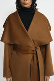 A close-up of a model wearing a camel brown wrap coat with a wide shawl collar and matching waist tie, layered over a black turtleneck.
