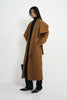 A model wearing a long camel brown wrap coat over a black turtleneck, black wide-leg trousers, and black pointed-toe heels, standing against a plain white studio background.