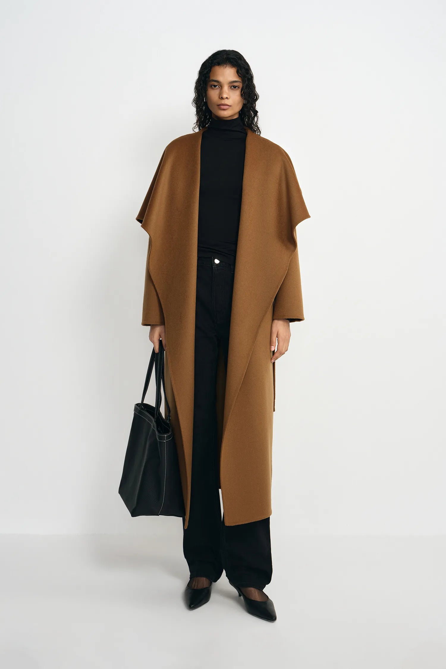 A model stands against a plain light gray studio background wearing a long camel-brown open-front coat over a black turtleneck, black wide-leg pants, and black pointed shoes, while holding a black tote bag.