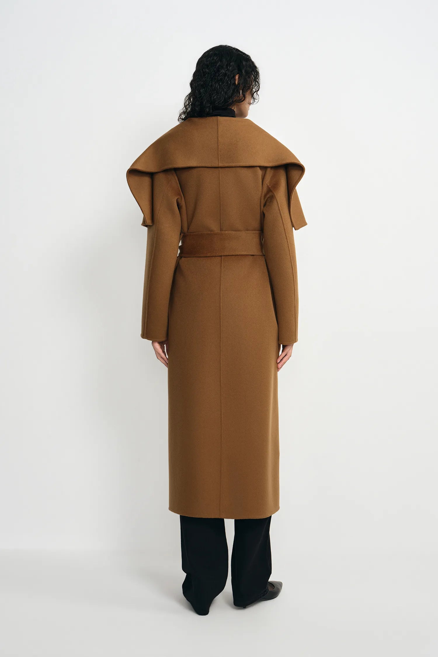A model viewed from the back wearing a camel brown long coat with a wide collar, a matching belt, and black trousers against a plain white studio background.