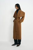 A woman with curly hair wearing a camel wool wrap coat with an oversized draped collar and self-tie belt, layered over a black turtleneck and black trousers, styled with black kitten-heel pumps, against a plain white background.