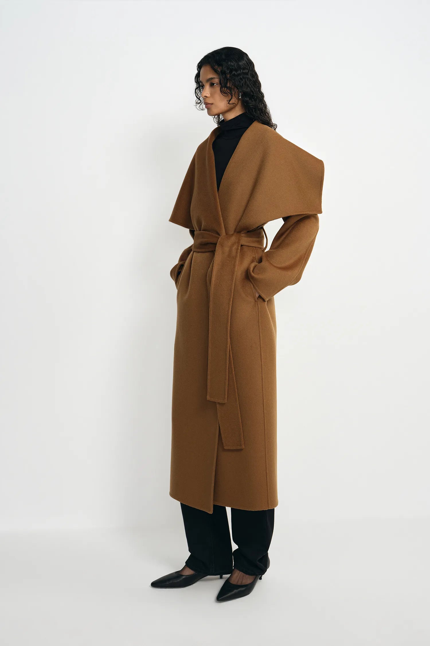 A woman with curly hair wearing a camel wool wrap coat with an oversized draped collar and self-tie belt, layered over a black turtleneck and black trousers, styled with black kitten-heel pumps, against a plain white background.