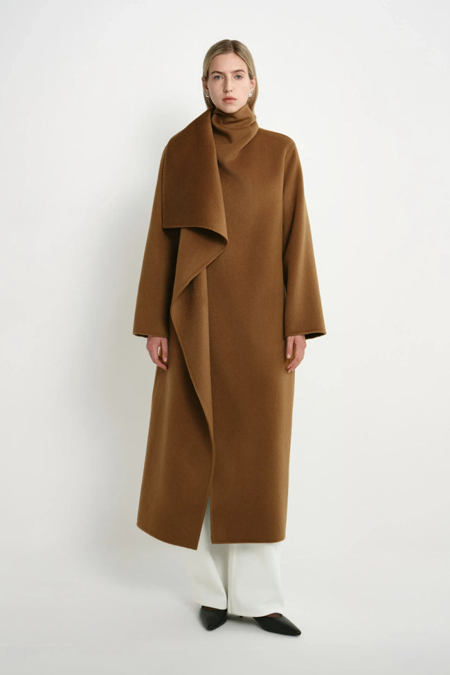 A model wearing a long, camel-brown wrap coat with a high draped collar, paired with white wide-leg trousers and black pointed shoes, standing against a plain white studio background.