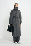 A model wearing a heather-grey wool-cashmere coat with the collar fastened high and waist belted, paired with charcoal trousers, white shoes and a black bag.