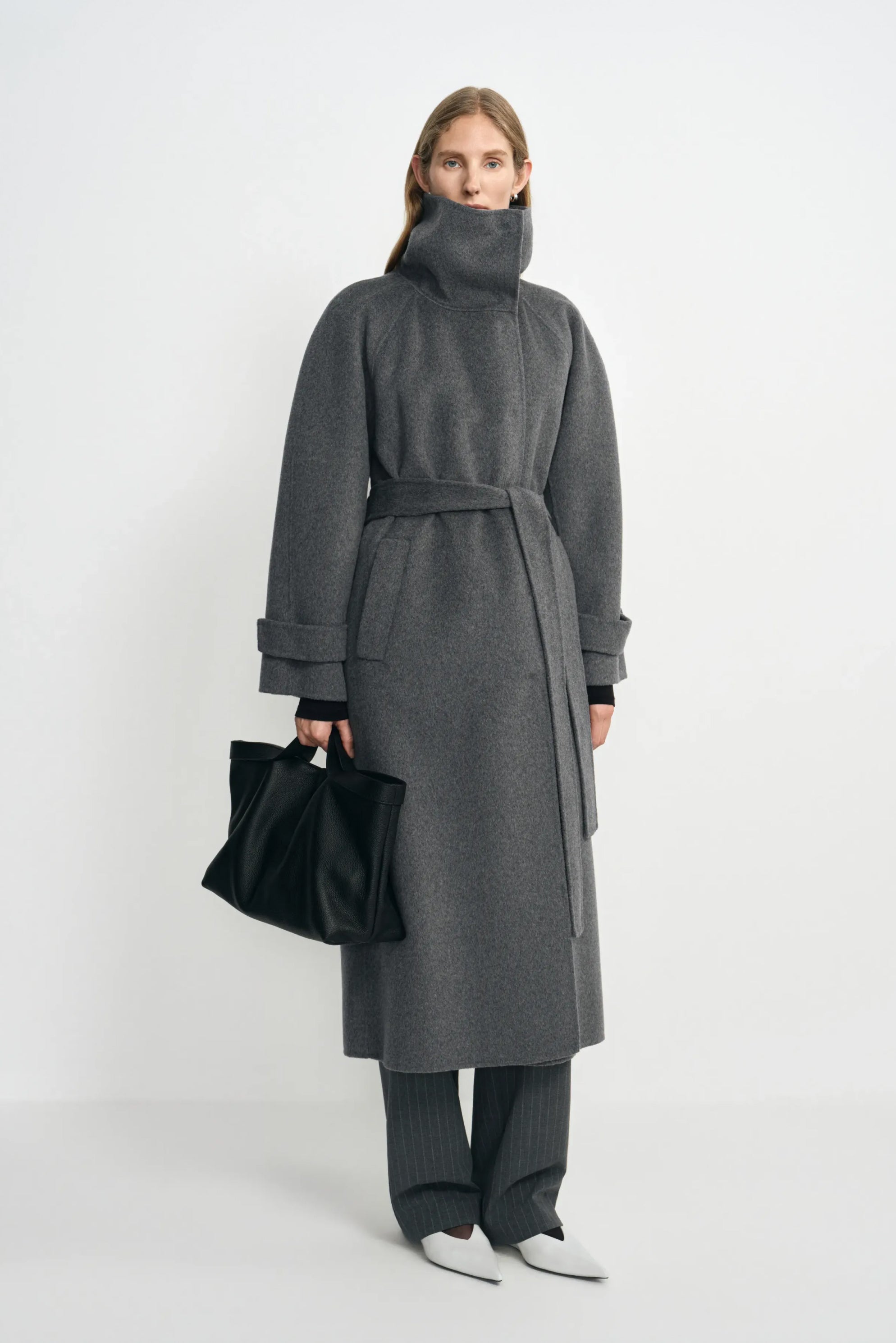 A model wearing a heather-grey wool-cashmere coat with the collar fastened high and waist belted, paired with charcoal trousers, white shoes and a black bag.