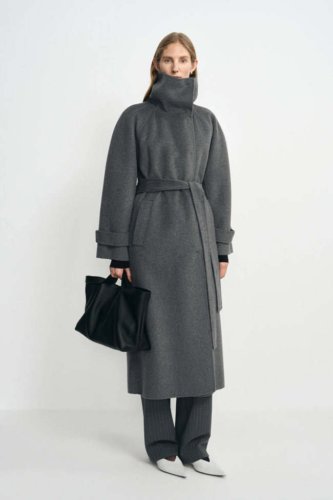 A model wearing a heather-grey wool-cashmere coat with the collar fastened high and waist belted, paired with charcoal trousers, white shoes and a black bag.