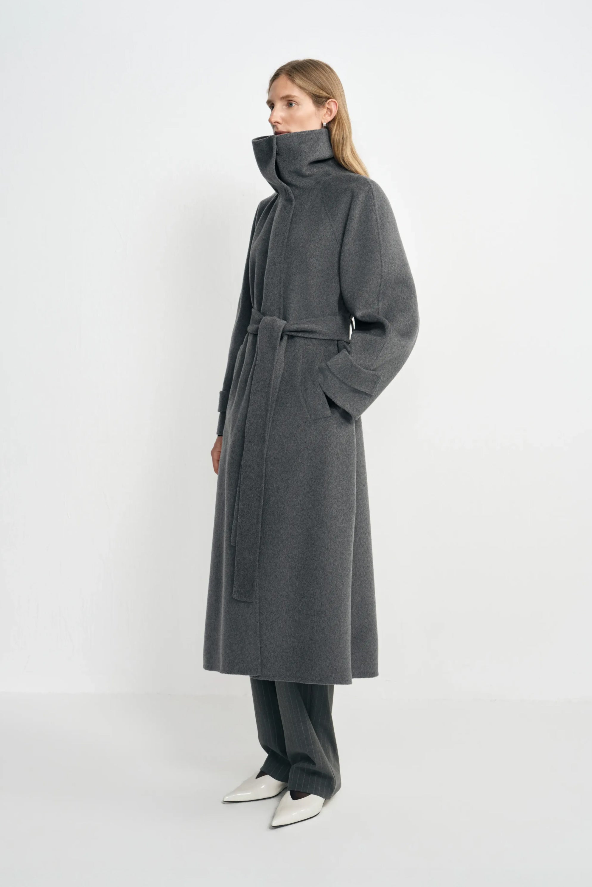 Side view of a model in a heather-grey coat with a high collar, belted waist and folded cuffs, worn over charcoal trousers with pointed white shoes.