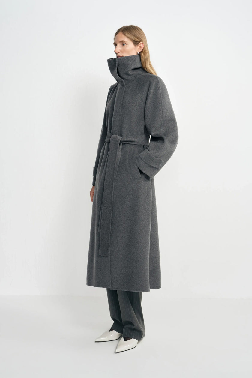 Side view of a model in a heather-grey coat with a high collar, belted waist and folded cuffs, worn over charcoal trousers with pointed white shoes.