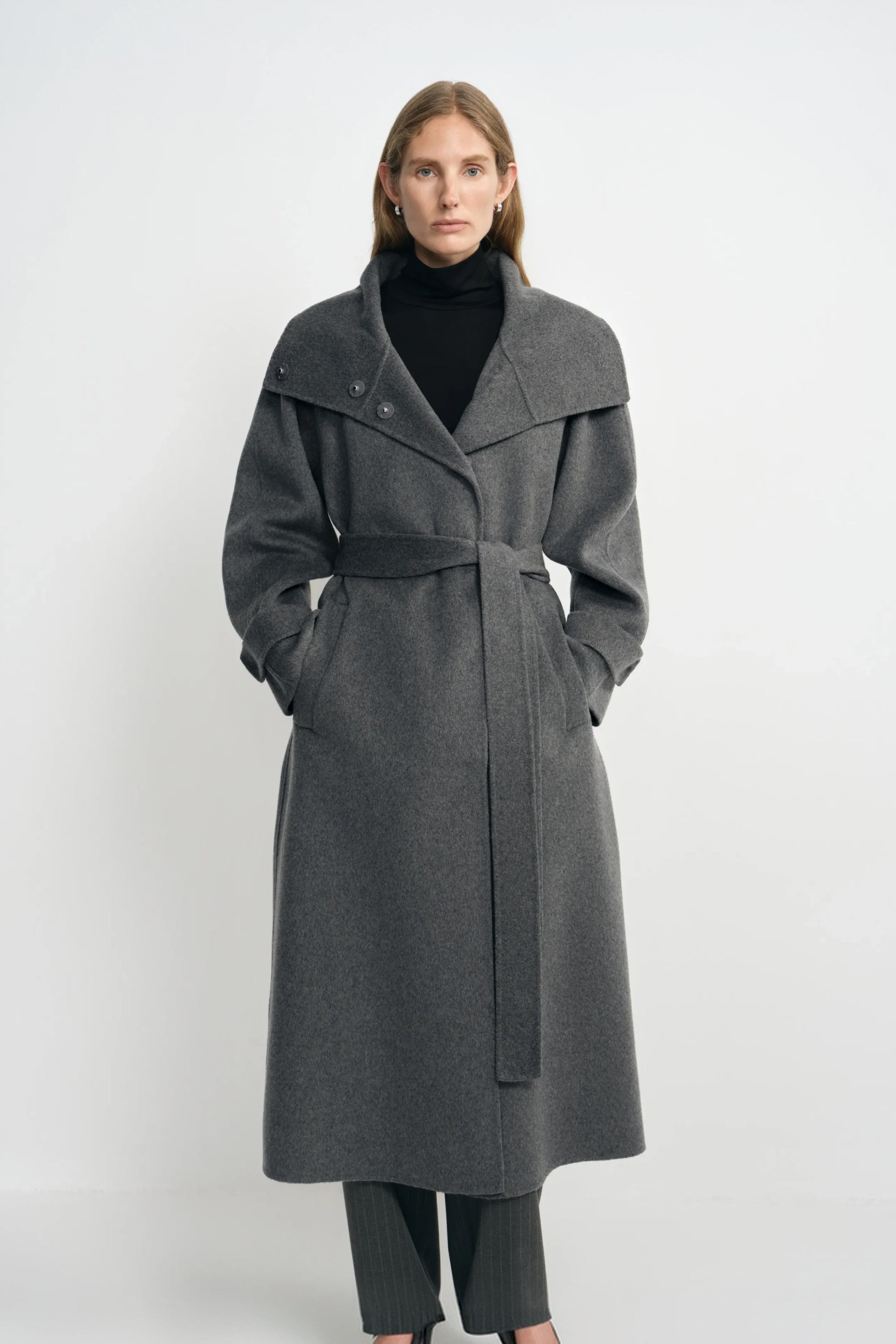 Front view of a model in a heather-grey coat with the collar folded wide and waist belted, hands in the pockets, worn over a black turtleneck.