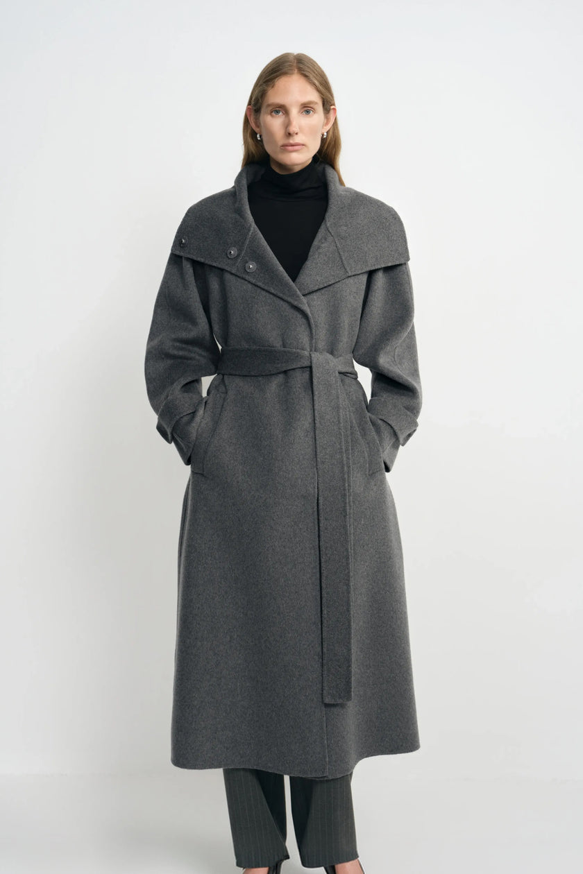 Front view of a model in a heather-grey coat with the collar folded wide and waist belted, hands in the pockets, worn over a black turtleneck.