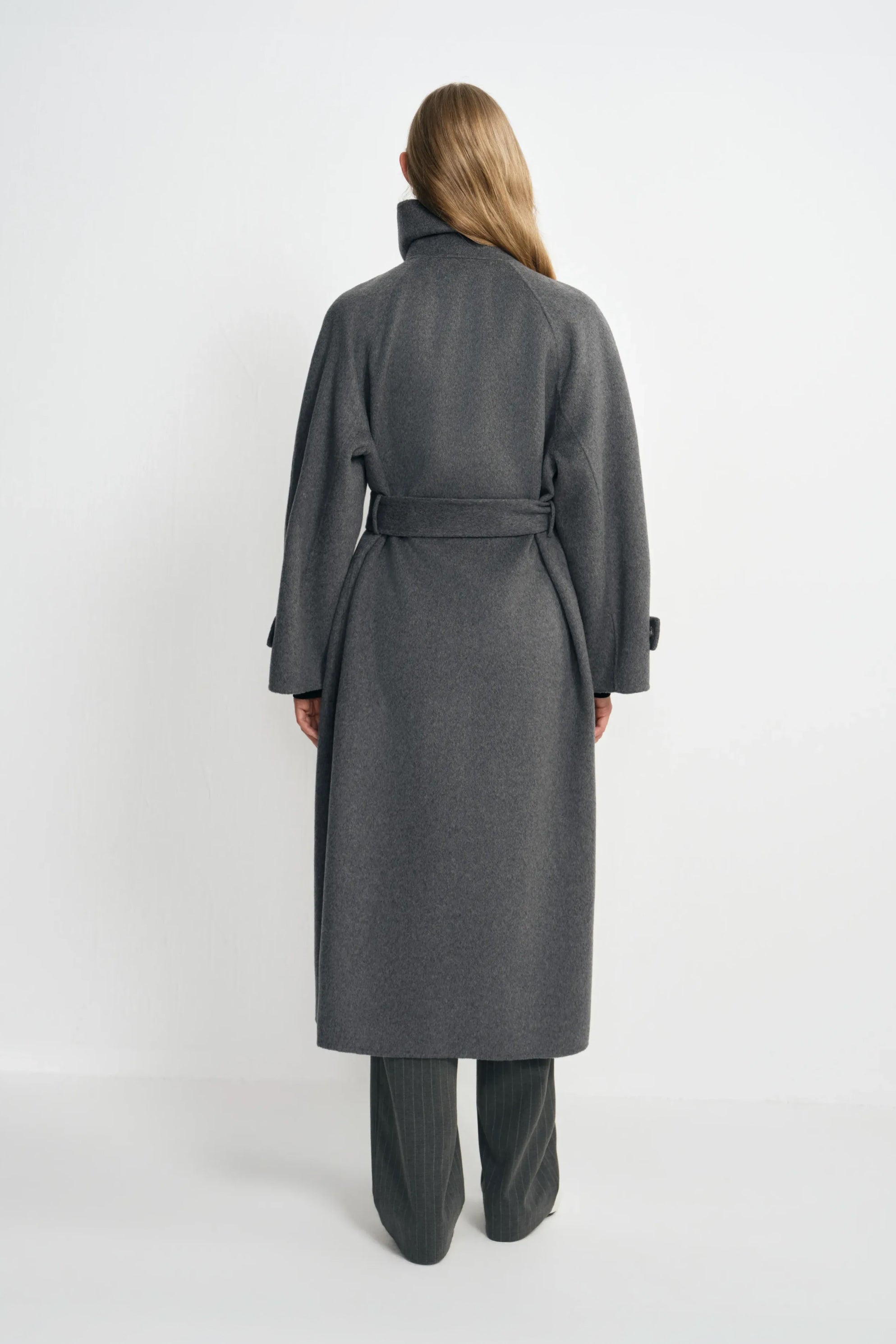 Back view of a model wearing a heather-grey wool-cashmere coat, with the belt drawing in the waist above a long hem and charcoal striped trousers.