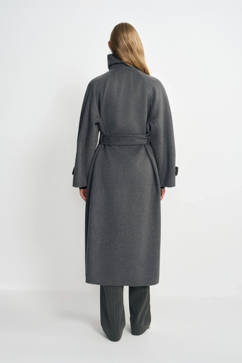 Back view of a model wearing a heather-grey wool-cashmere coat, with the belt drawing in the waist above a long hem and charcoal striped trousers.