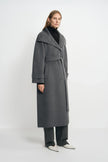 Three-quarter view of a model in a heather-grey belted coat with a wide open collar and turned cuffs, paired with charcoal trousers and white pointed shoes.