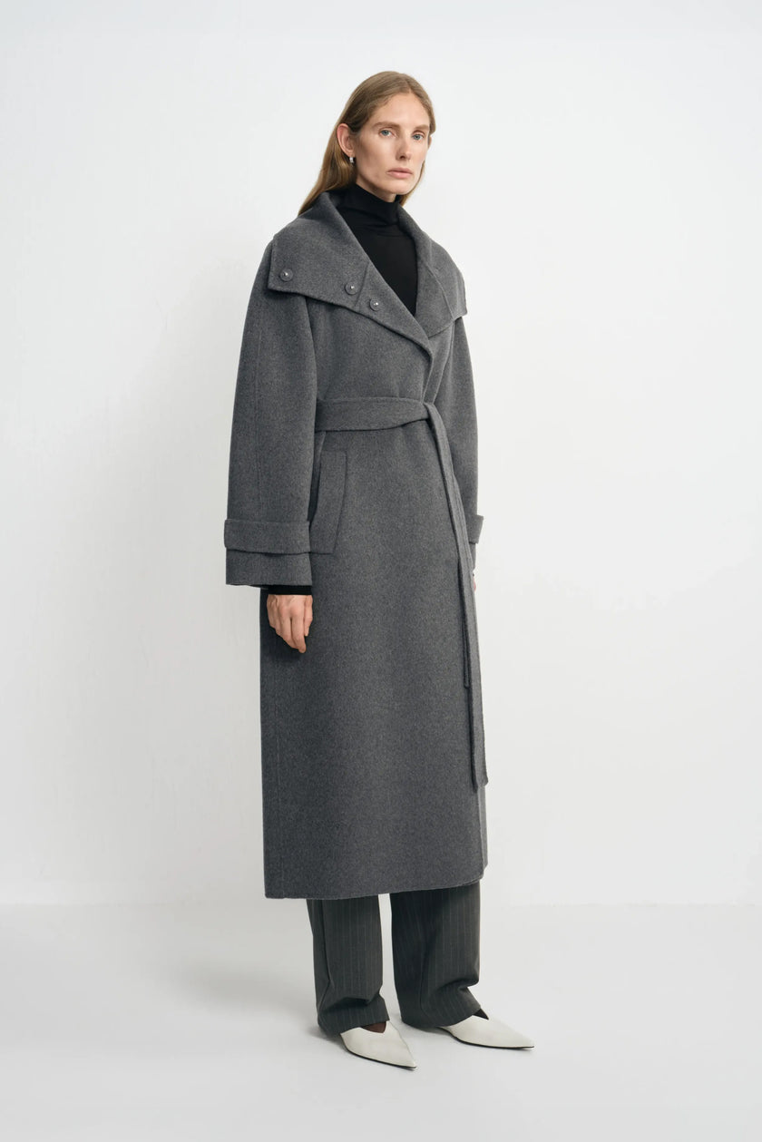 Three-quarter view of a model in a heather-grey belted coat with a wide open collar and turned cuffs, paired with charcoal trousers and white pointed shoes.
