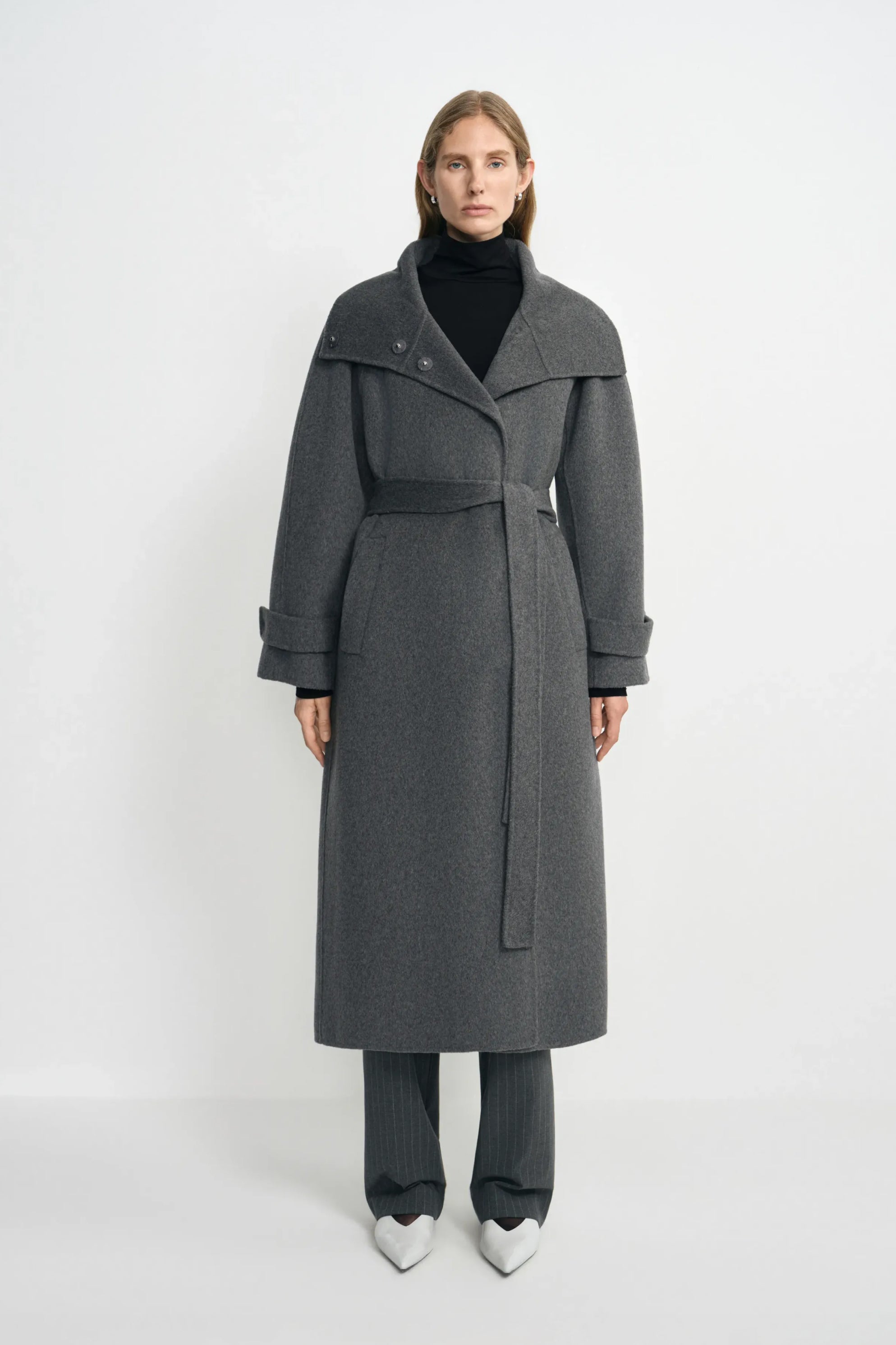 A model wearing a heather-grey wool-cashmere coat with a wide folded collar and tied waist, over a black turtleneck and striped trousers with white shoes.
