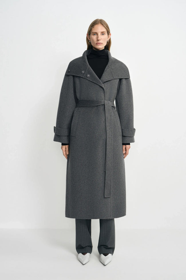 A model wearing a heather-grey wool-cashmere coat with a wide folded collar and tied waist, over a black turtleneck and striped trousers with white shoes.