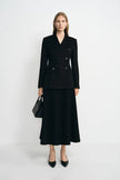 A woman stands against a light gray studio background wearing a black double-breasted blazer with a matching midi skirt, black heels, and carrying a black handbag.