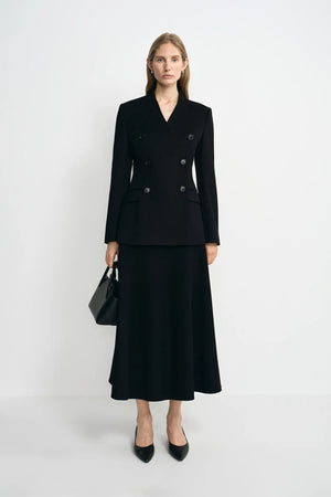 A woman stands against a light gray studio background wearing a black double-breasted blazer with a matching midi skirt, black heels, and carrying a black handbag.