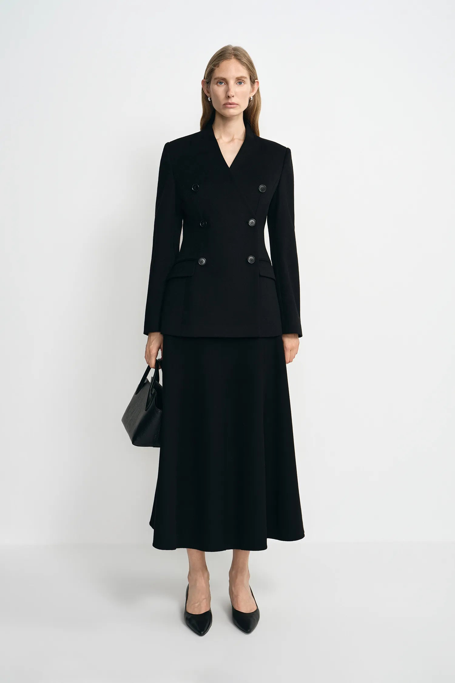 A woman stands against a light gray studio background wearing a black double-breasted blazer with a matching midi skirt, black heels, and carrying a black handbag.