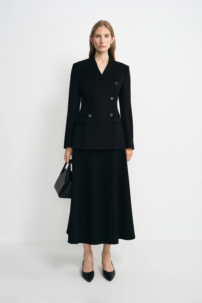 A woman stands against a light gray studio background wearing a black double-breasted blazer with a matching midi skirt, black heels, and carrying a black handbag.