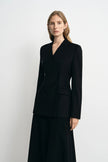 A woman modeling a black double-breasted blazer with a V-neckline and matching black skirt against a plain light gray studio background.