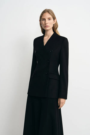 A woman modeling a black double-breasted blazer with a V-neckline and matching black skirt against a plain light gray studio background.