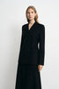 A woman modeling a black double-breasted blazer with a V-neckline and matching black skirt against a plain light gray studio background.