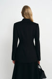 A woman shown from the back wearing a structured black blazer and black skirt, holding a black handbag against a plain light gray studio background.