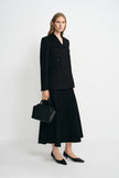 A woman modeling a black double-breasted blazer and matching midi skirt, holding a structured black handbag and wearing black pointed flats against a clean white studio background.