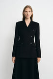 A woman modeling a black double-breasted blazer with structured shoulders and a matching black skirt against a plain light gray studio background.