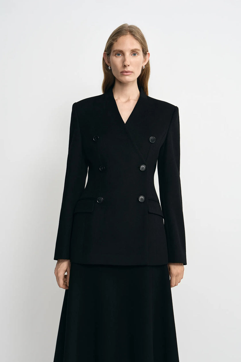 A woman modeling a black double-breasted blazer with structured shoulders and a matching black skirt against a plain light gray studio background.