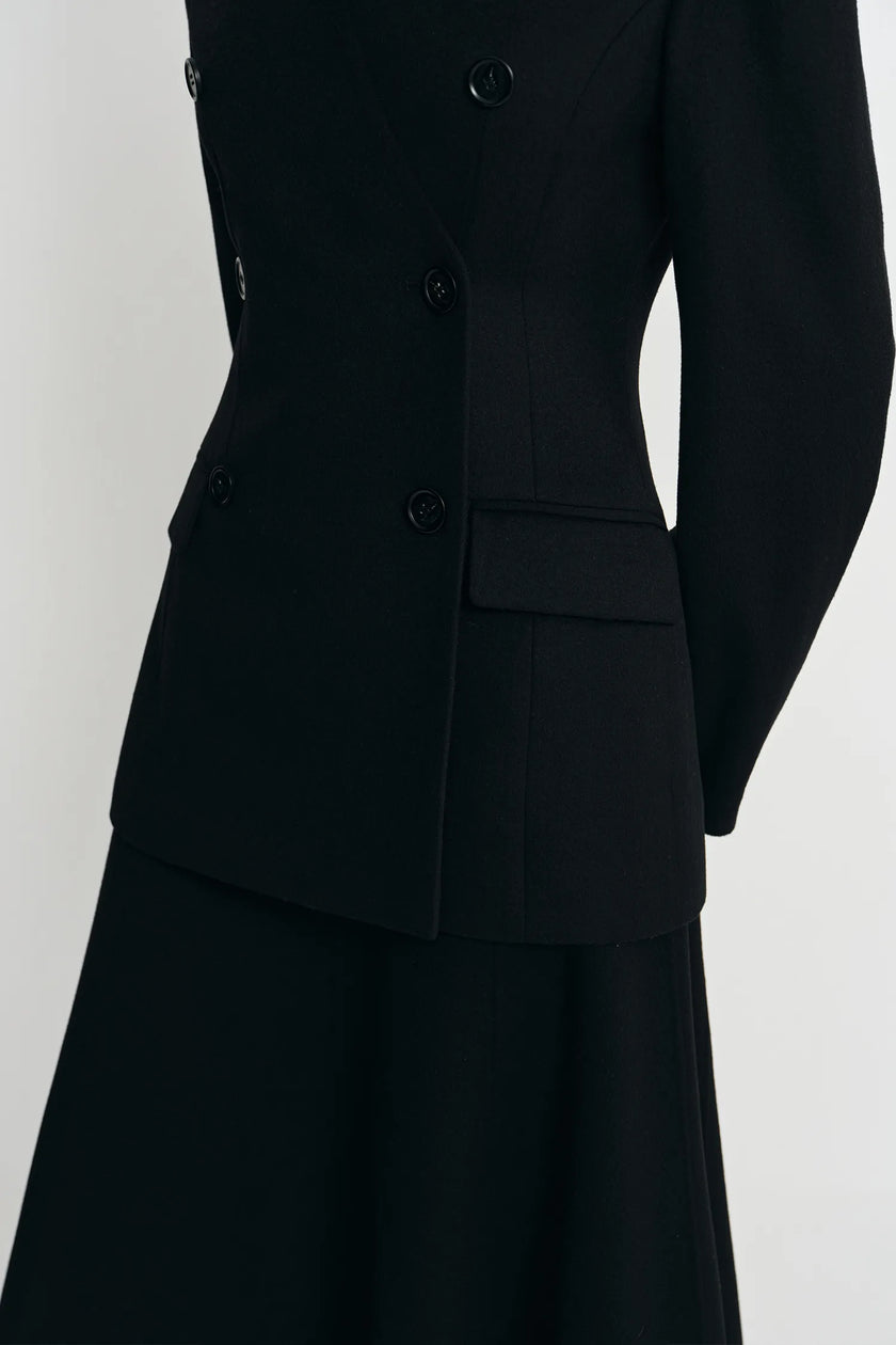 A close-up of a black double-breasted blazer with structured shoulders and a matching black skirt, shown against a plain light gray background.