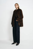 A model standing at an angle in a brown collarless faux fur coat with full sleeves, worn open over a black top and dark jeans with black pointed shoes.